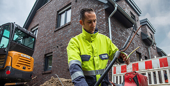 Man in high-visibility clothing holding several exposed power cables at a construction site in front of a residential building.