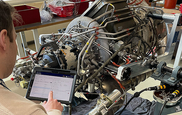 A person documents maintenance data for an Auxiliary Power Unit (APU) on a tablet. The auxiliary engine is mounted on a test stand in a workshop environment.  