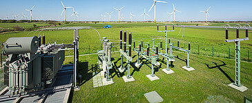Umspannwerk auf einer grünen Wiese mit Windenergieanlagen im Hintergrund