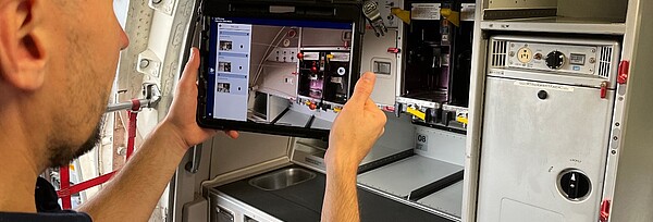 A person holds a tablet showing an image of the interior of a technical cabin. The cabin contains switches, sockets, and metal compartments.