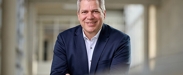 Mann mit kurzen grauen Haaren, braunen Augen, dunkelbauen Jackett und weißem Hemd