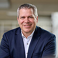 Mann mit kurzen grauen Haaren, braunen Augen, dunkelbauen Jackett und weißem Hemd