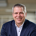 Mann mit kurzen grauen Haaren, braunen Augen, dunkelbauen Jackett und weißem Hemd