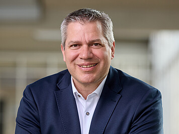 Mann mit kurzen grauen Haaren, braunen Augen, dunkelbauen Jackett und weißem Hemd