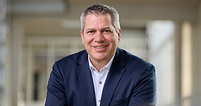 Mann mit kurzen grauen Haaren, braunen Augen, dunkelbauen Jackett und weißem Hemd