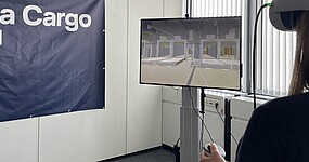 Person nutzt eine VR-Brille zur Simulation einer Lager- oder Logistikumgebung. Auf einem Monitor wird die virtuelle Umgebung angezeigt, im Hintergrund ist ein Lufthansa Cargo eVo-Banner zu sehen.
