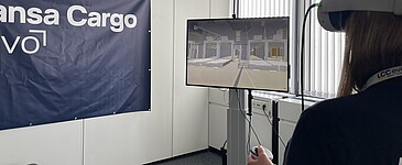 Person nutzt eine VR-Brille zur Simulation einer Lager- oder Logistikumgebung. Auf einem Monitor wird die virtuelle Umgebung angezeigt, im Hintergrund ist ein Lufthansa Cargo eVo-Banner zu sehen.