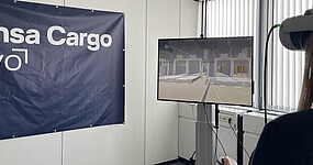 A person is using VR goggles to simulate a warehouse or logistics environment. The virtual environment is displayed on a monitor, and a Lufthansa Cargo eVo banner is visible in the background.