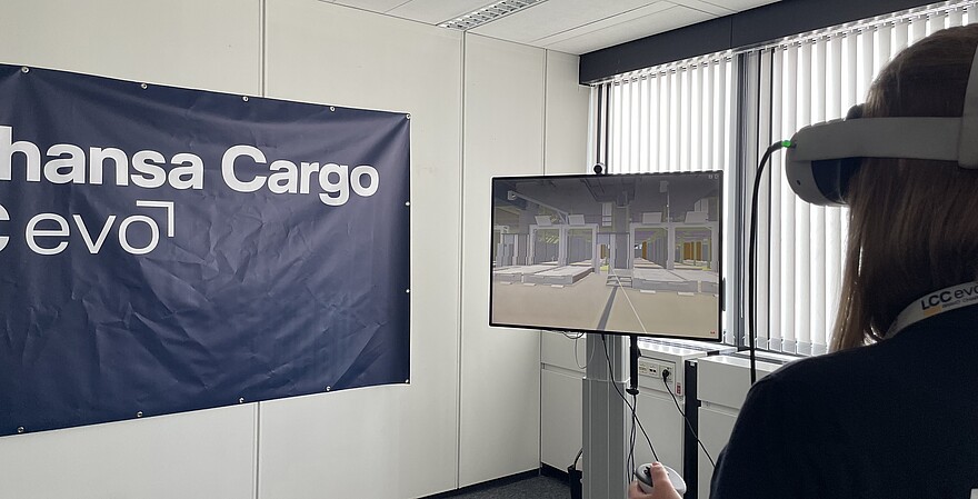 Person nutzt eine VR-Brille zur Simulation einer Lager- oder Logistikumgebung. Auf einem Monitor wird die virtuelle Umgebung angezeigt, im Hintergrund ist ein Lufthansa Cargo eVo-Banner zu sehen.