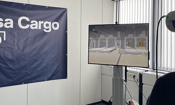 A person is using VR goggles to simulate a warehouse or logistics environment. The virtual environment is displayed on a monitor, and a Lufthansa Cargo eVo banner is visible in the background.
