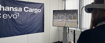 A person is using VR goggles to simulate a warehouse or logistics environment. The virtual environment is displayed on a monitor, and a Lufthansa Cargo eVo banner is visible in the background.