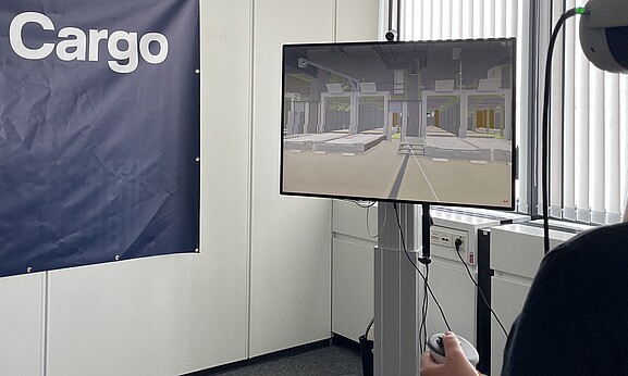 Person nutzt eine VR-Brille zur Simulation einer Lager- oder Logistikumgebung. Auf einem Monitor wird die virtuelle Umgebung angezeigt, im Hintergrund ist ein Lufthansa Cargo eVo-Banner zu sehen.