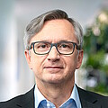 Mann mit kurzen grauen Haaren, braunen Augen, grau umrandeter Brille, dunklem Jackett und hellblauem Hemd