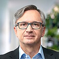 Mann mit kurzen grauen Haaren, braunen Augen, grau umrandeter Brille, dunklem Jackett und hellblauem Hemd
