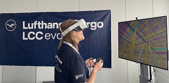 Person mit VR-Brille und Controllern betrachtet eine digitale 3D-Modellierung auf einem Monitor vor einem Lufthansa Cargo LCC eVo-Hintergrund.