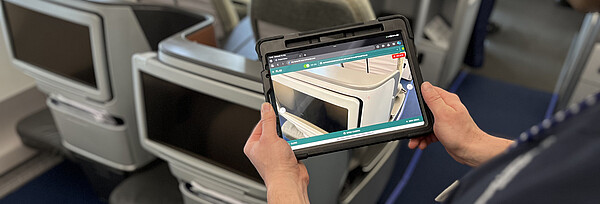Ein Mann steht am Sitzplatz in der Flugzeugkabine und hält ein Tablet in den Händen. Auf dem Tablet sieht man den Sitzplatz wie durch eine Kamera.