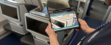 Ein Mann steht am Sitzplatz in der Flugzeugkabine und hält ein Tablet in den Händen. Auf dem Tablet sieht man den Sitzplatz wie durch eine Kamera.