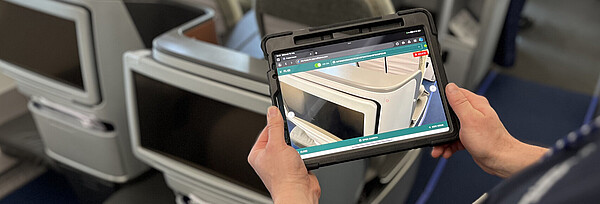 A man is standing at a seat in the airplane cabin, holding a tablet in his hands. On the tablet, you can see the seat as if through a camera.