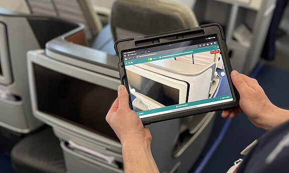 Ein Mann steht am Sitzplatz in der Flugzeugkabine und hält ein Tablet in den Händen. Auf dem Tablet sieht man den Sitzplatz wie durch eine Kamera.