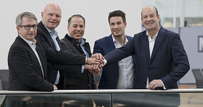 Five men in dark suits and white shirts hold their hands stacked on top of each other and smile at the camera. They are in the waiting area of an airport.